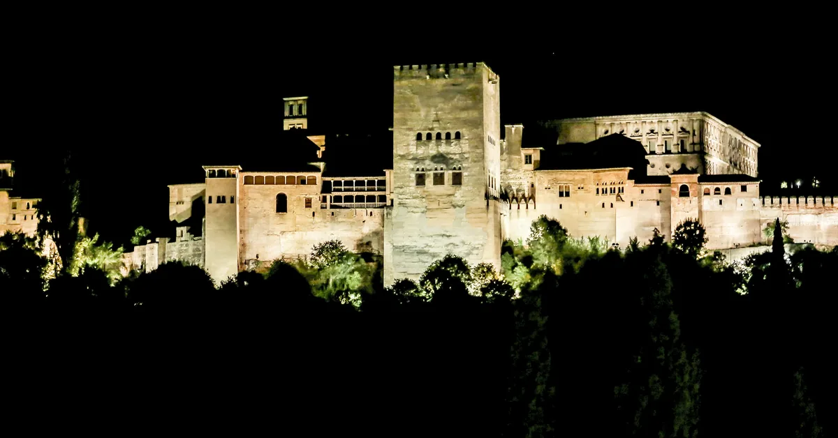 The Alhambra's walls and towers floodlit above dark trees in Granada at night